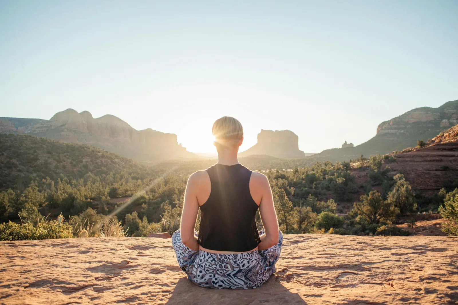 Back view of anonymous fit woman in comfortable summer wear sitting in Padmasana and meditating on sunlit cliff at sunrise