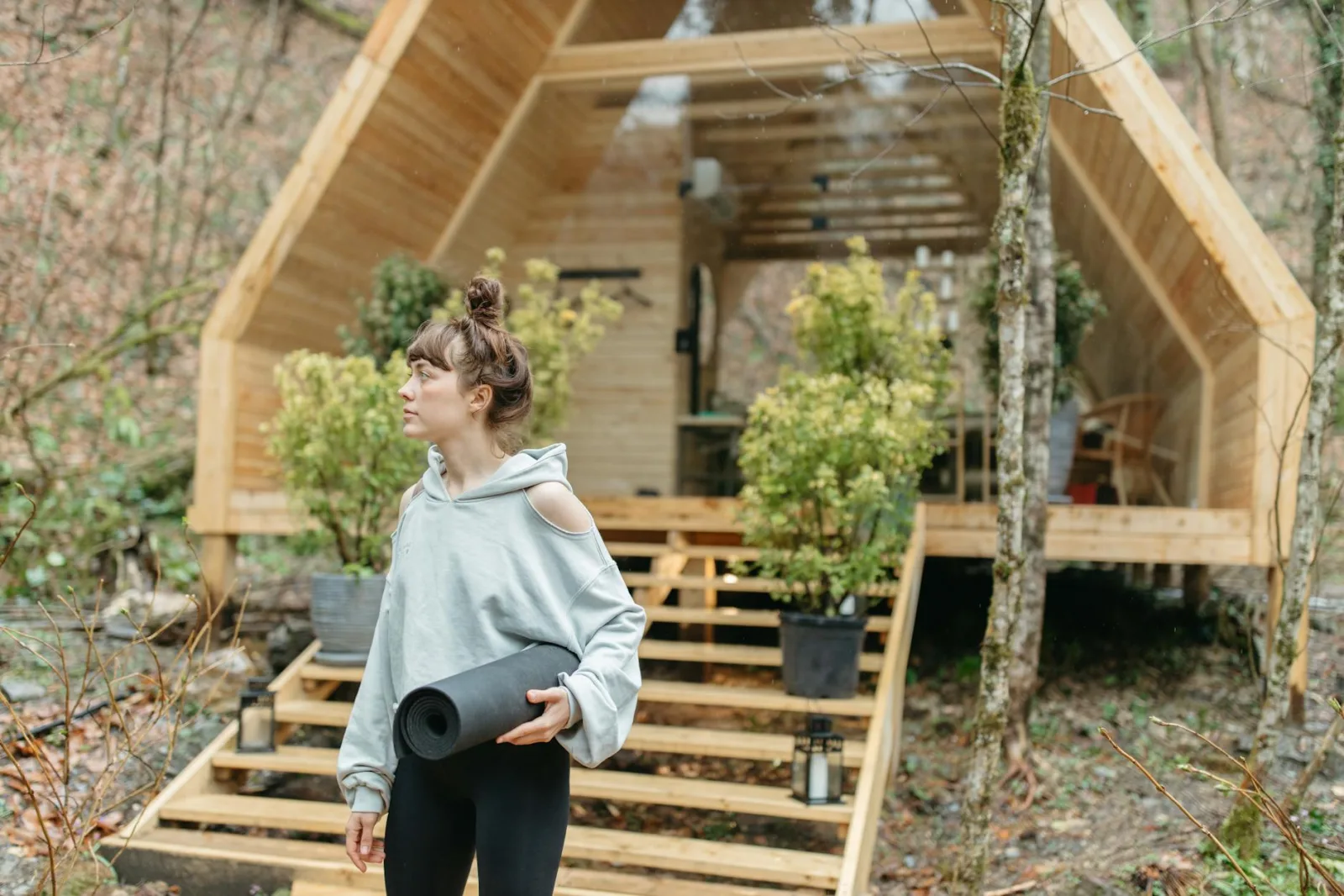 Woman holding a yoga mat outside a wooden cabin in a forest, ready for a wellness retreat.