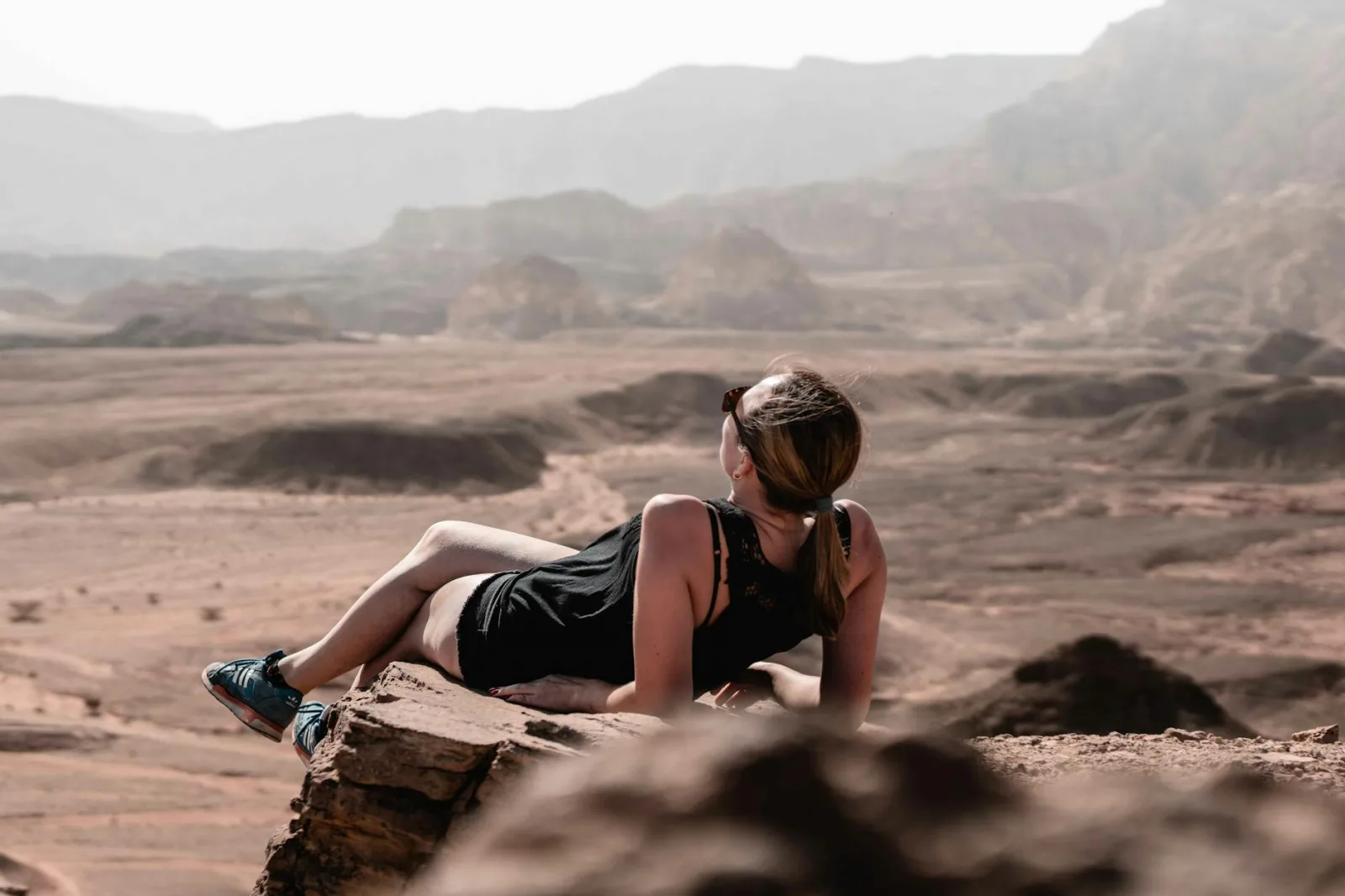 Woman in sunglasses relaxing on rocky terrain with expansive desert view.