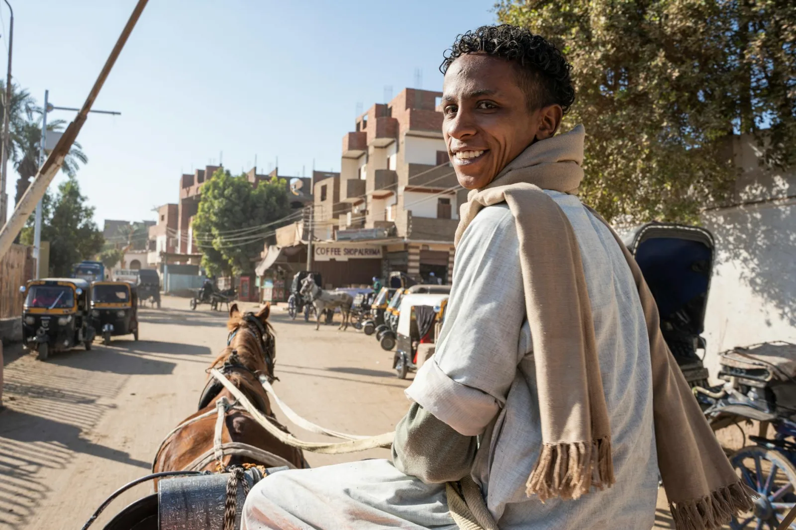 A man smiling while riding a horse-drawn carriage on a vibrant street lined with vehicles and shops.
