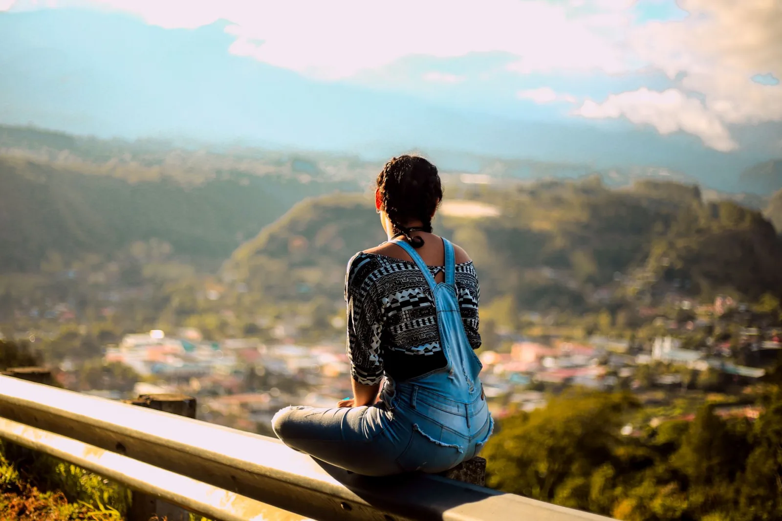 A woman sits on a guardrail enjoying a breathtaking mountain view.