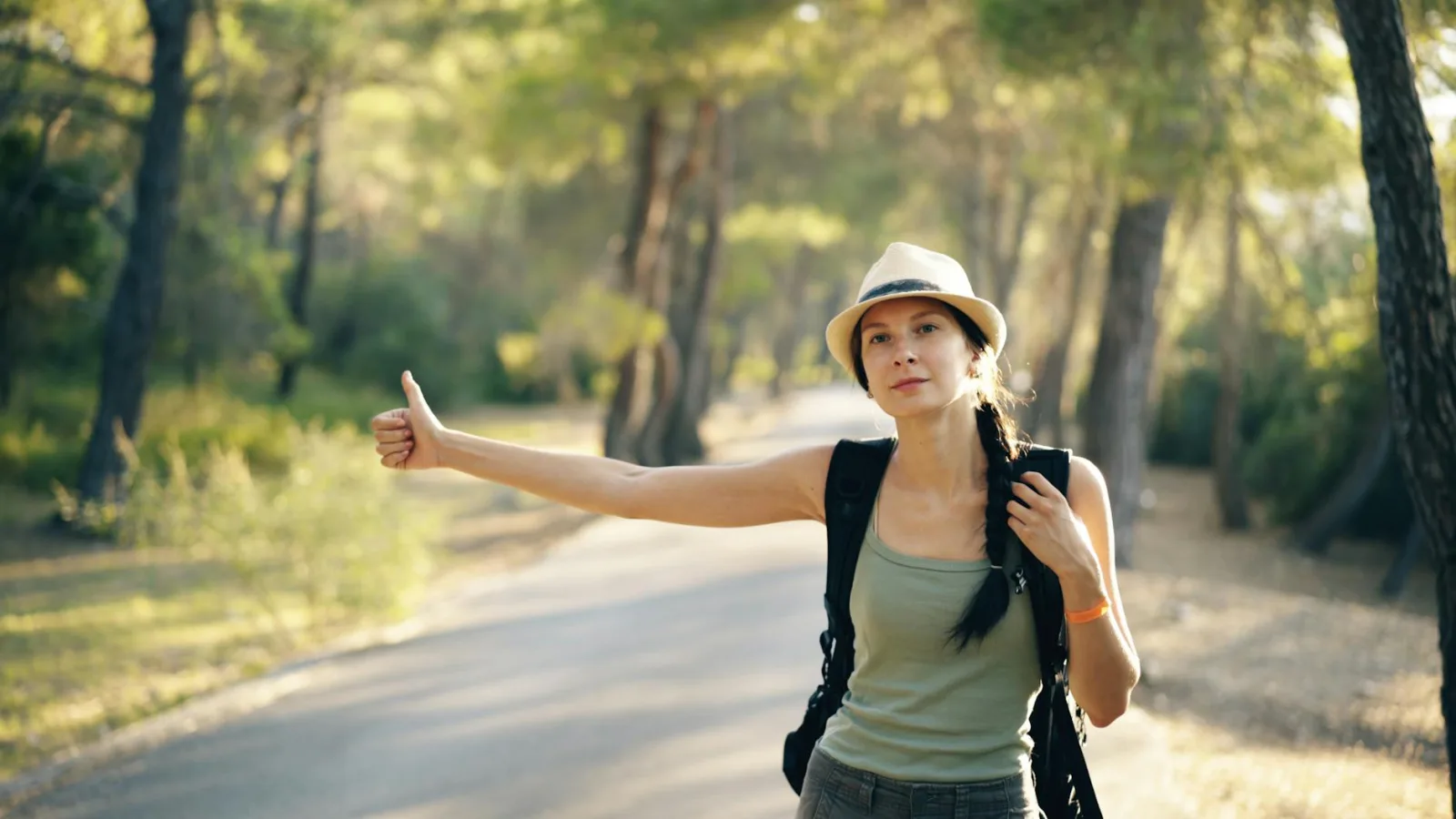 Woman in hat hitchhiking on a scenic forest road with sunlight filtering through trees.