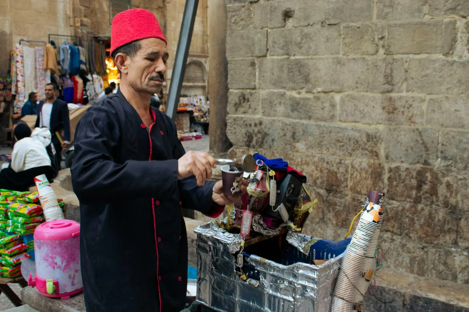 A street vendor in a red fez prepares a traditional drink at a bustling market.