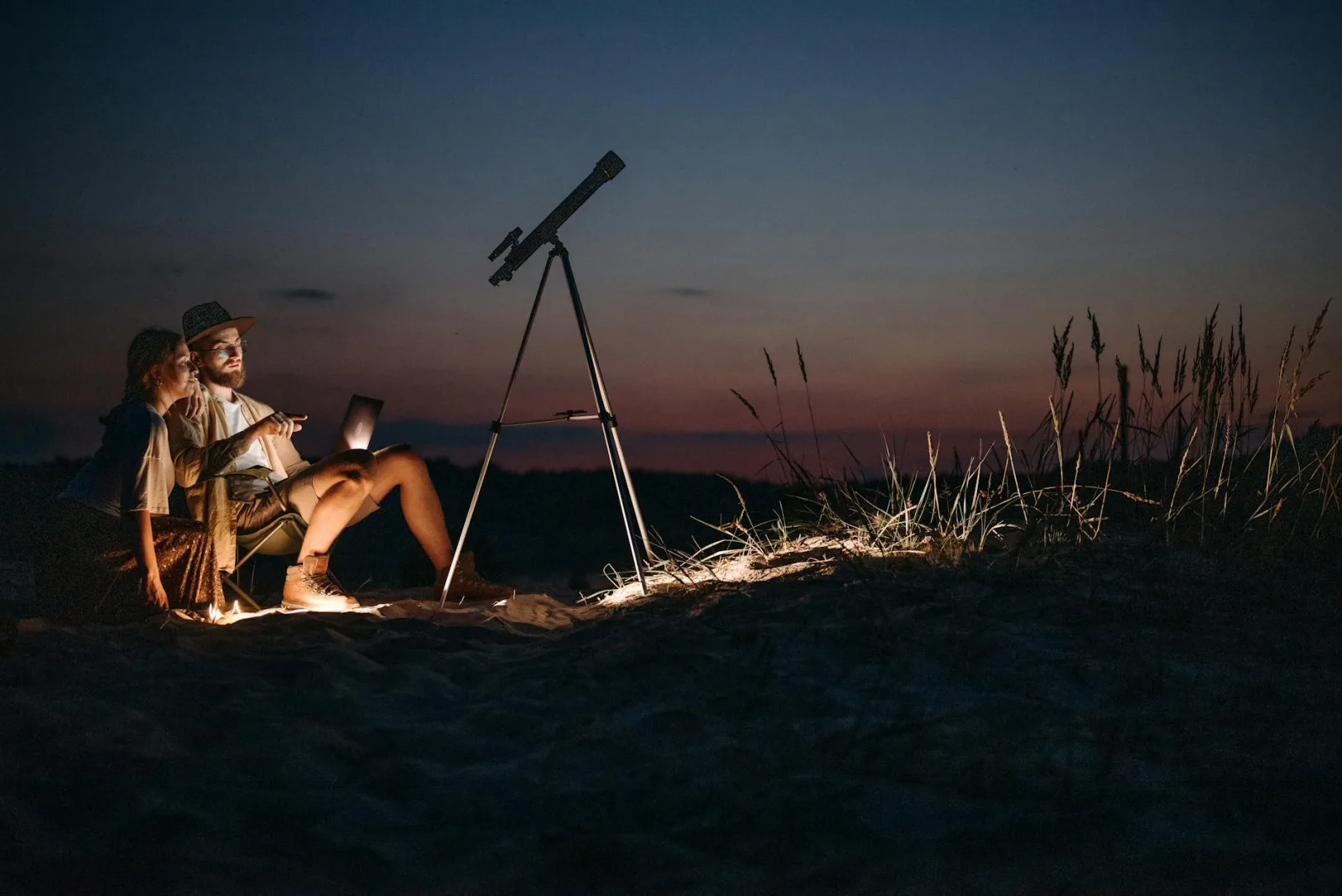 A couple enjoys stargazing with a telescope and laptop in the wilderness during a twilight camping adventure.