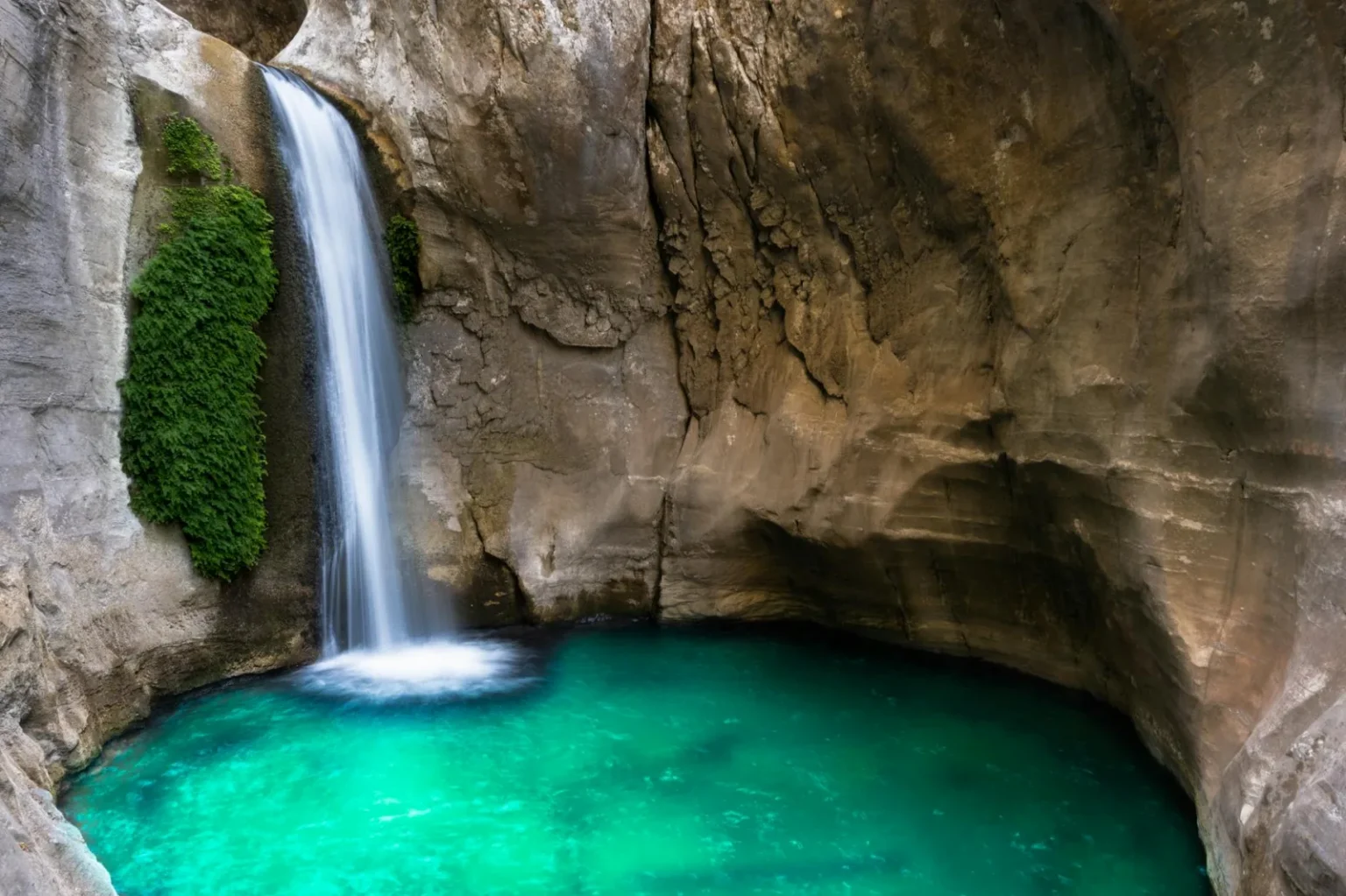 A beautiful waterfall flows into an emerald pool inside a rocky cave, creating a serene natural scene.