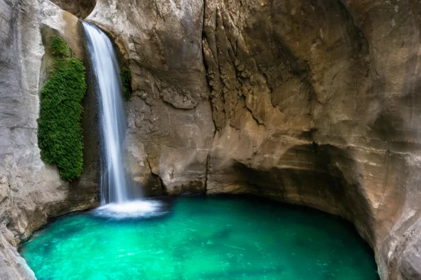 A beautiful waterfall flows into an emerald pool inside a rocky cave, creating a serene natural scene.