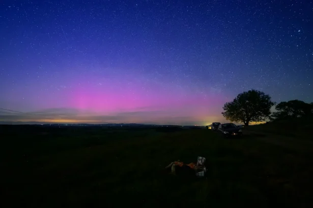 Vibrant night sky with pink aurora and stars over a serene meadow with cars parked under a tree.
