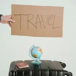 A hand holds a travel sign above a globe and suitcase, symbolizing wanderlust.