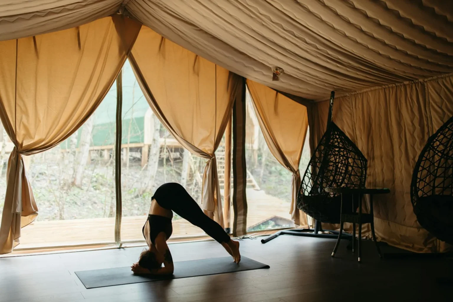 Adult woman performing yoga pose inside a tent with nature view, promoting serenity and wellness.