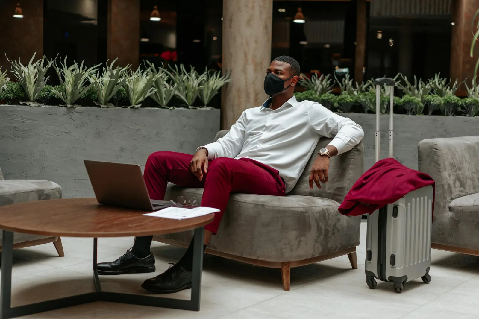 Man wearing facemask, sitting in hotel lobby with laptop and suitcase, adapting to new normal.