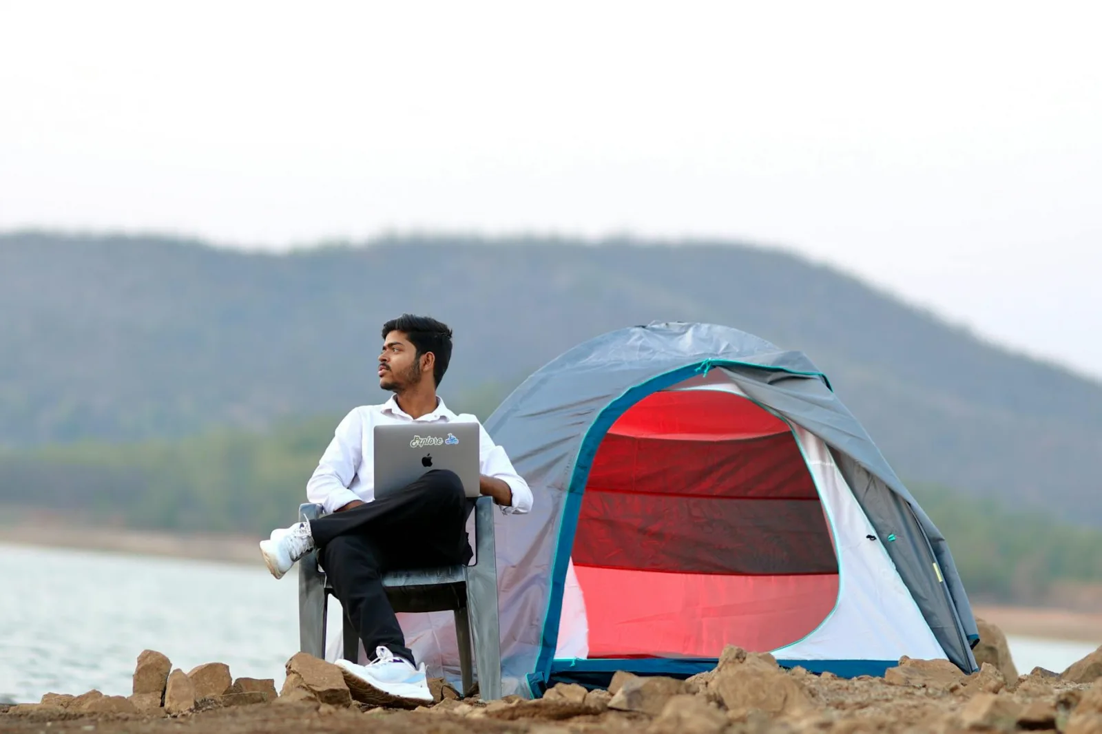 Man working on laptop by a lakeside tent, blending nature with remote work.