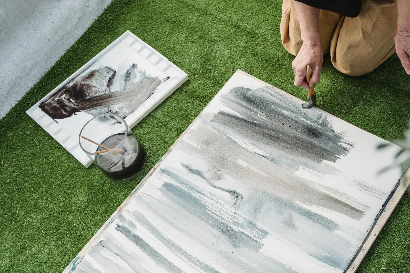 Person painting abstract art on canvas with brush outdoors on grass.