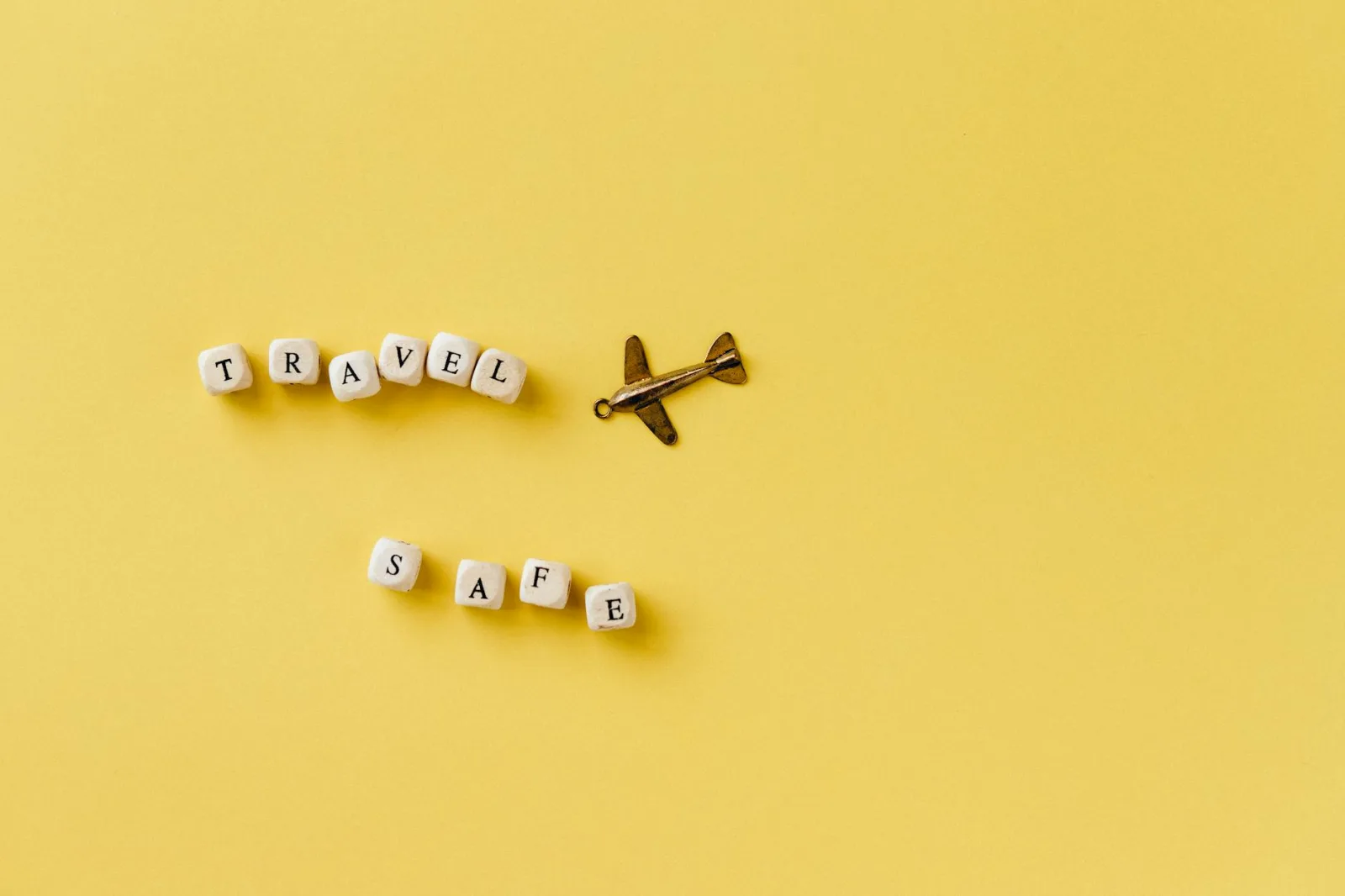 Flat lay of 'Travel Safe' text with airplane pendant on yellow background, evoking travel and safety concepts.