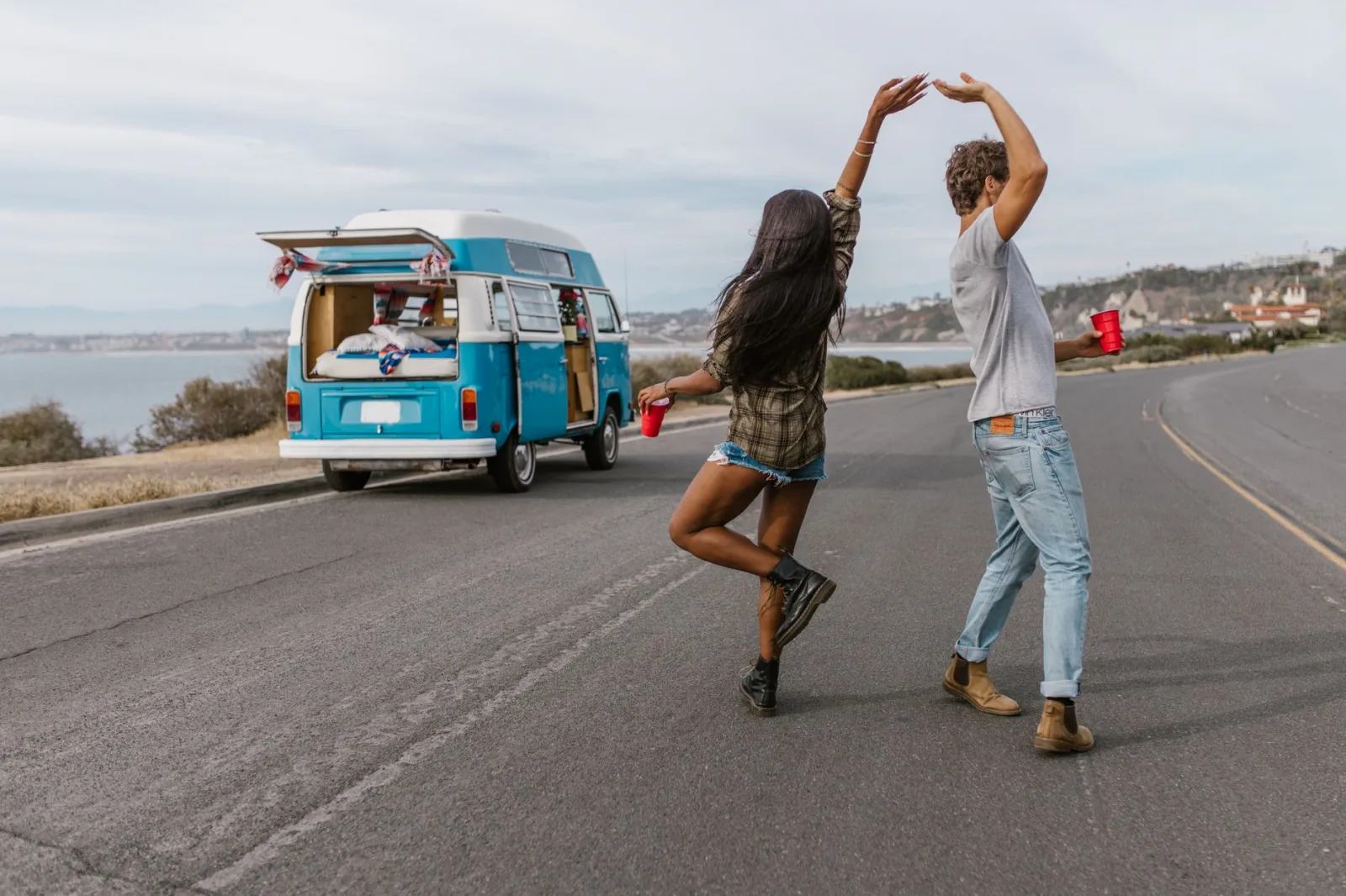 A carefree couple dancing by a vintage van on a scenic coastal road.