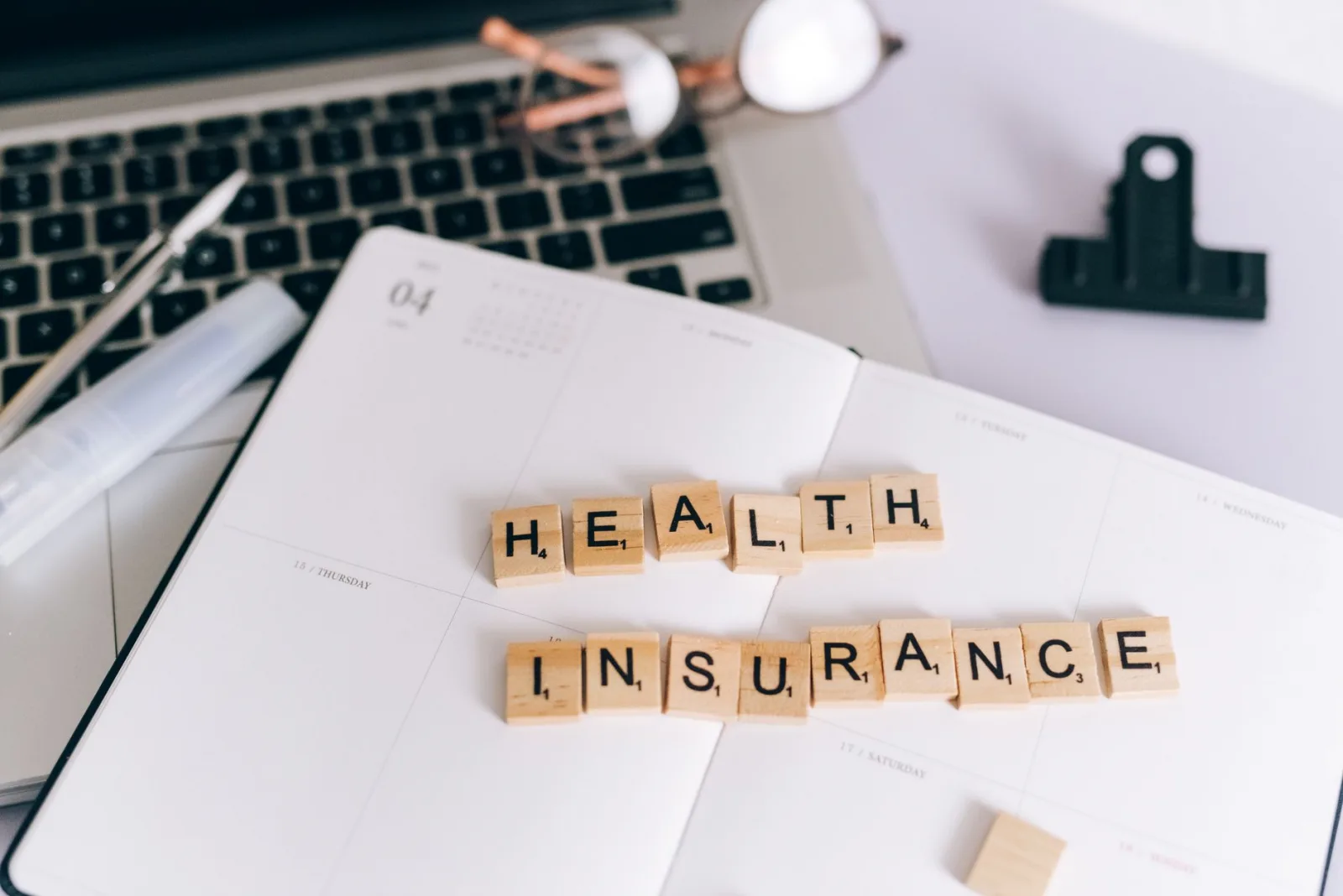 Close-up of Scrabble tiles spelling 'Health Insurance' on a planner with a laptop.