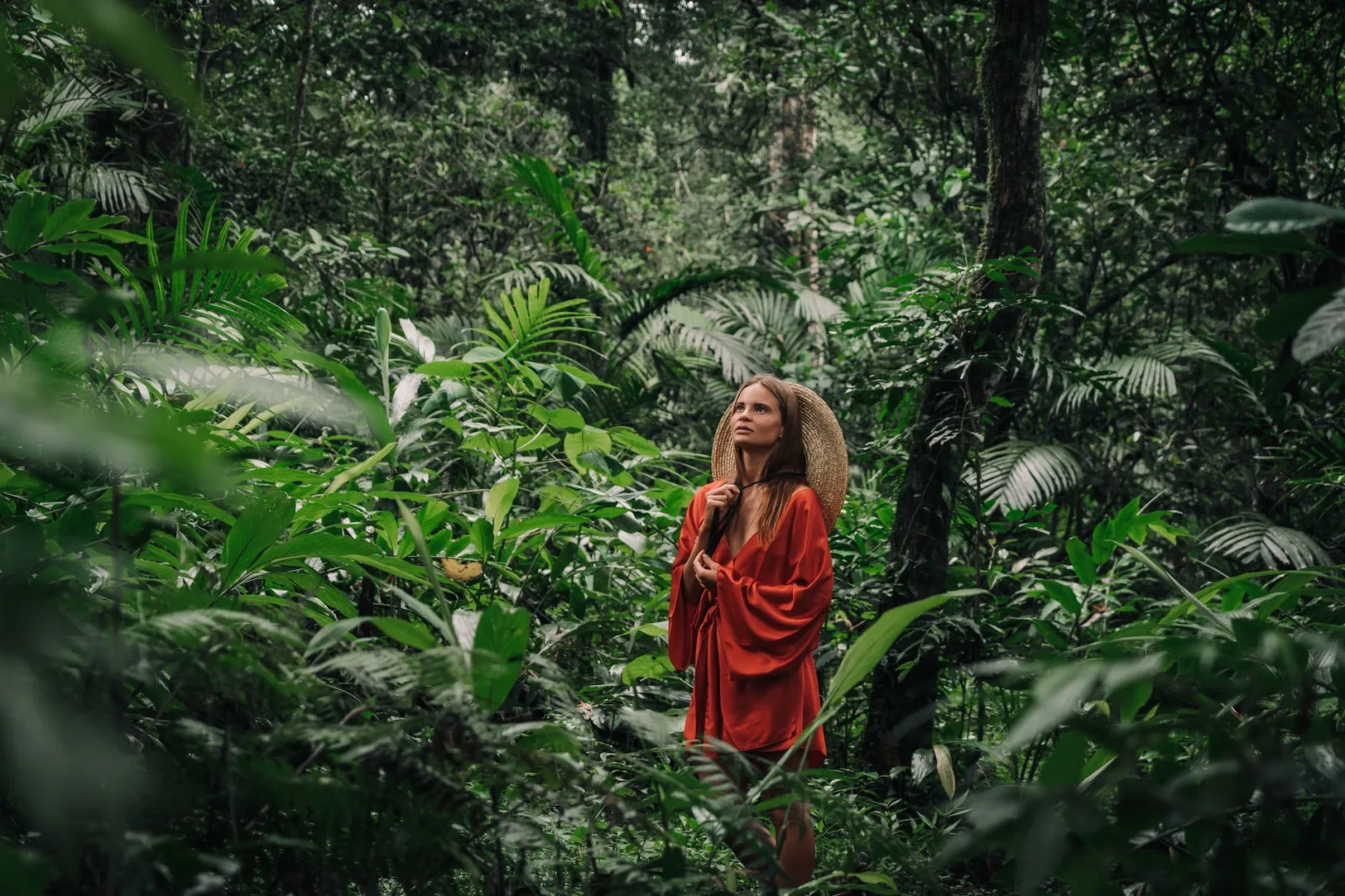 A woman stands in a vibrant rainforest wearing a red dress, embracing nature's serenity.