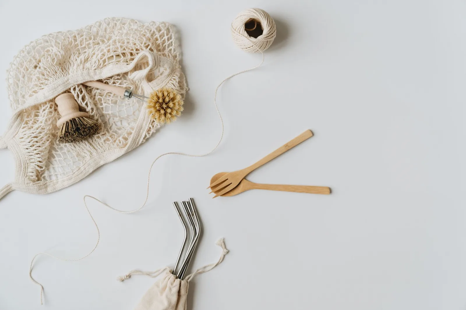 Flat lay of sustainable kitchen items including net bag, wooden utensils, and metal straws on a white surface.