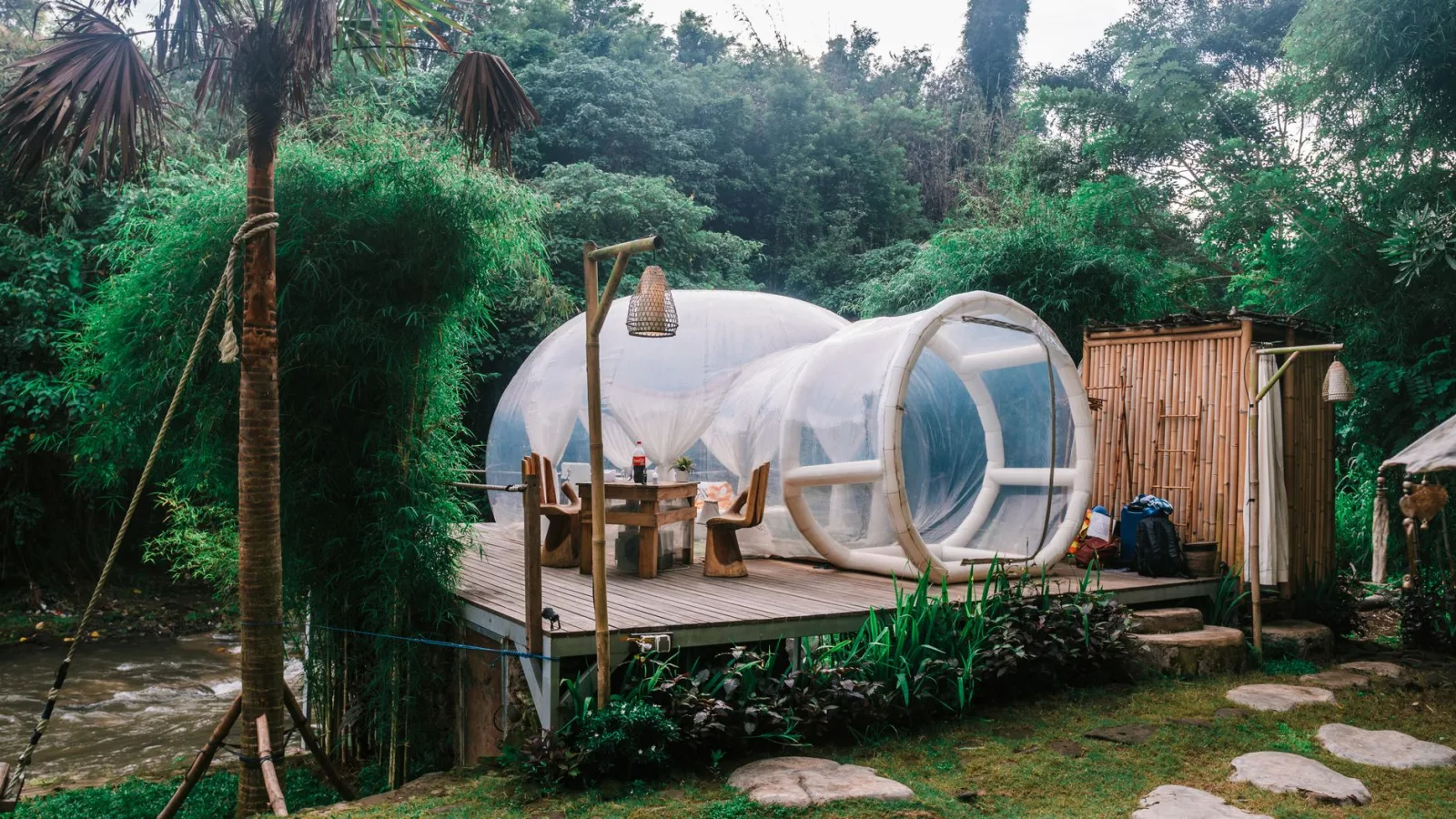 Cozy transparent bubble tent located on wooden platform near dining table in tropical forest