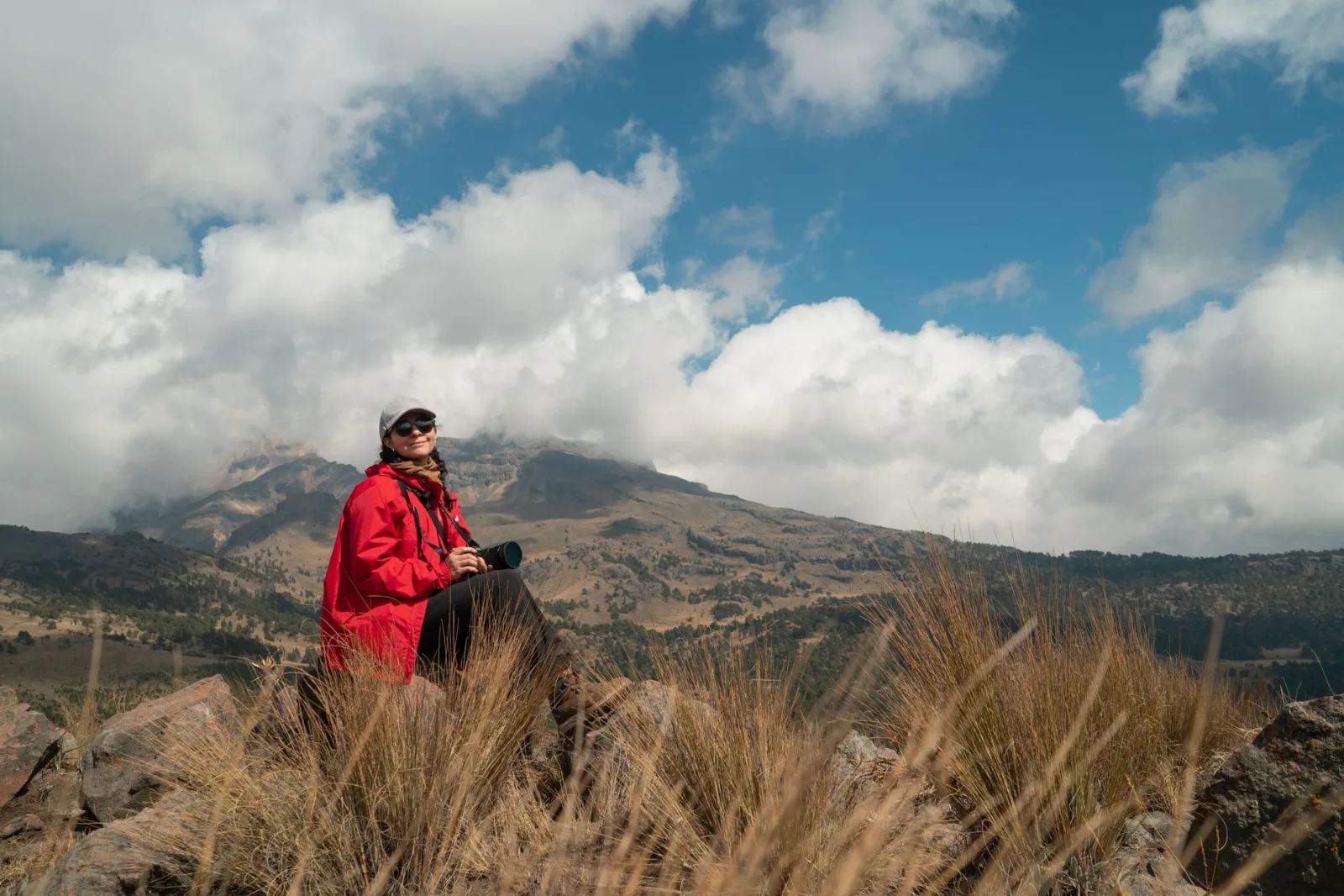 Woman resting on rocky hill with camera, enjoying mountainous landscape under a cloudy sky.