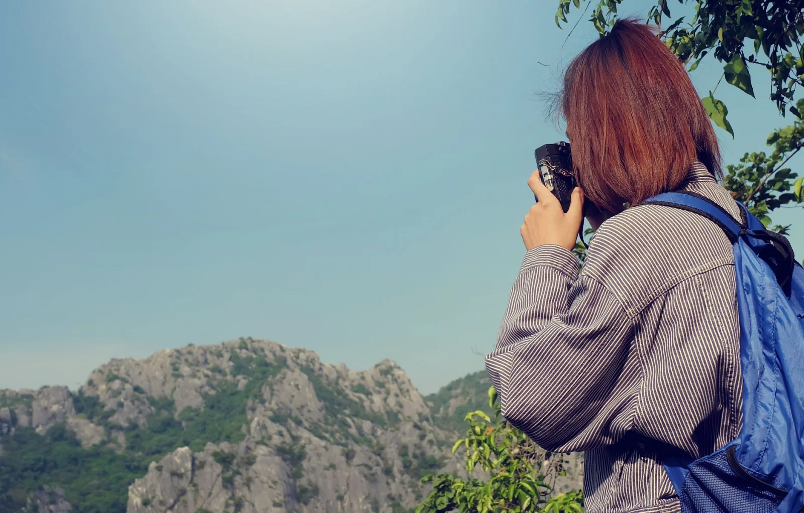 Woman photographing a scenic mountain view with clear sky and rocky peaks.