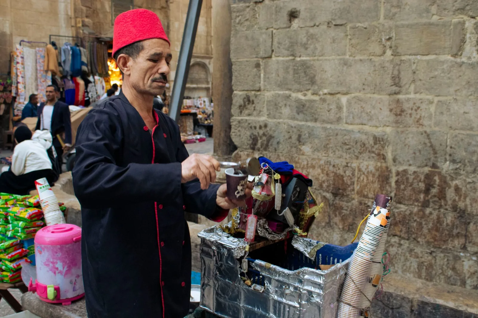 A street vendor in a red fez prepares a traditional drink at a bustling market.
