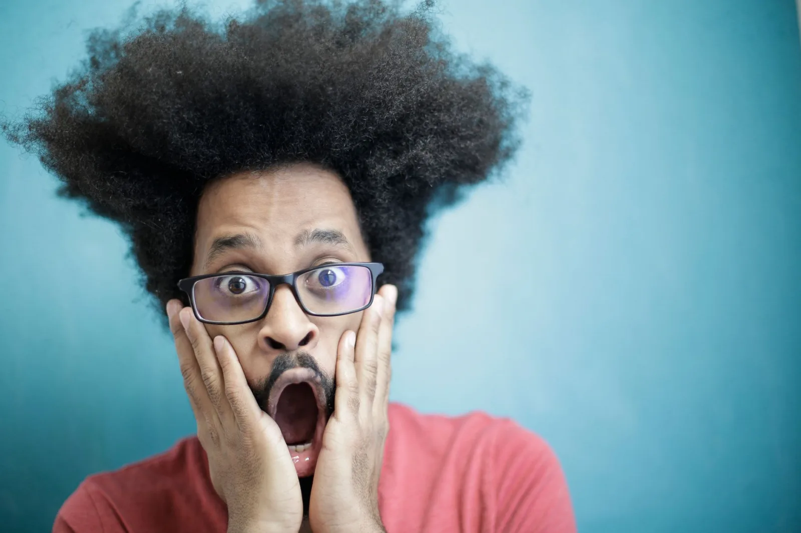 Close-up portrait of a surprised man with afro hairstyle and glasses against a blue background.
