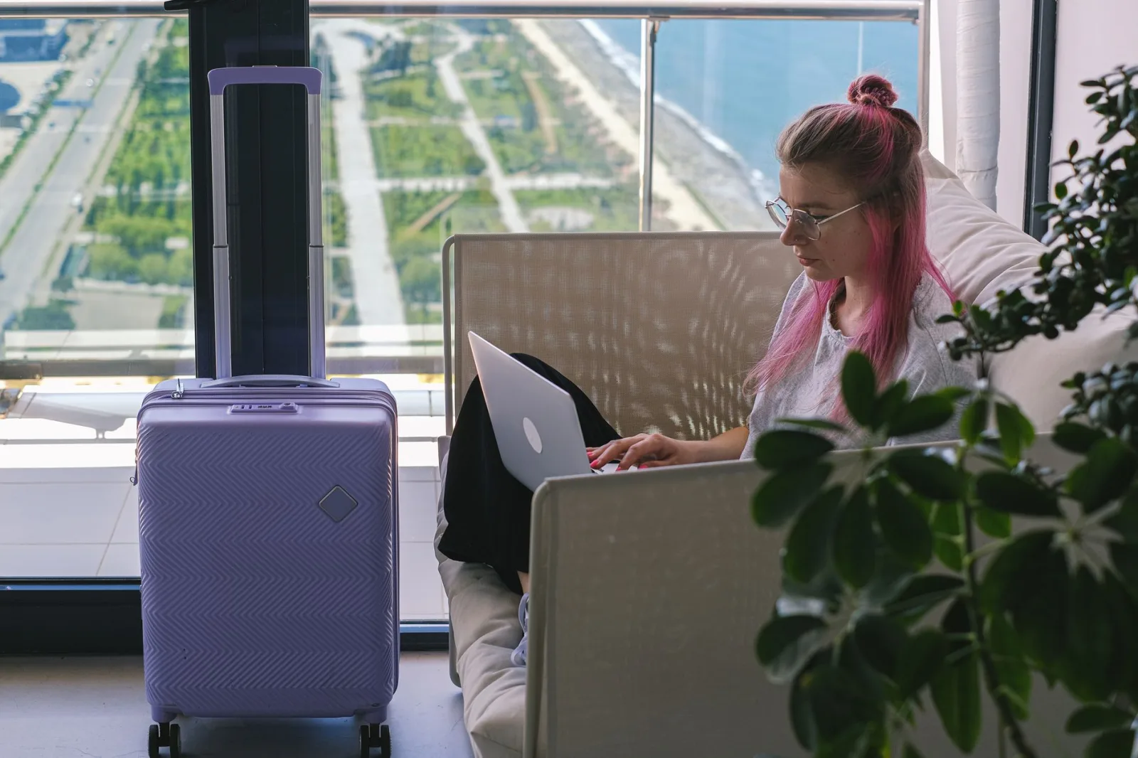 Woman with pink hair working on laptop in hotel room overlooking scenic landscape. Travel and freelance concept.