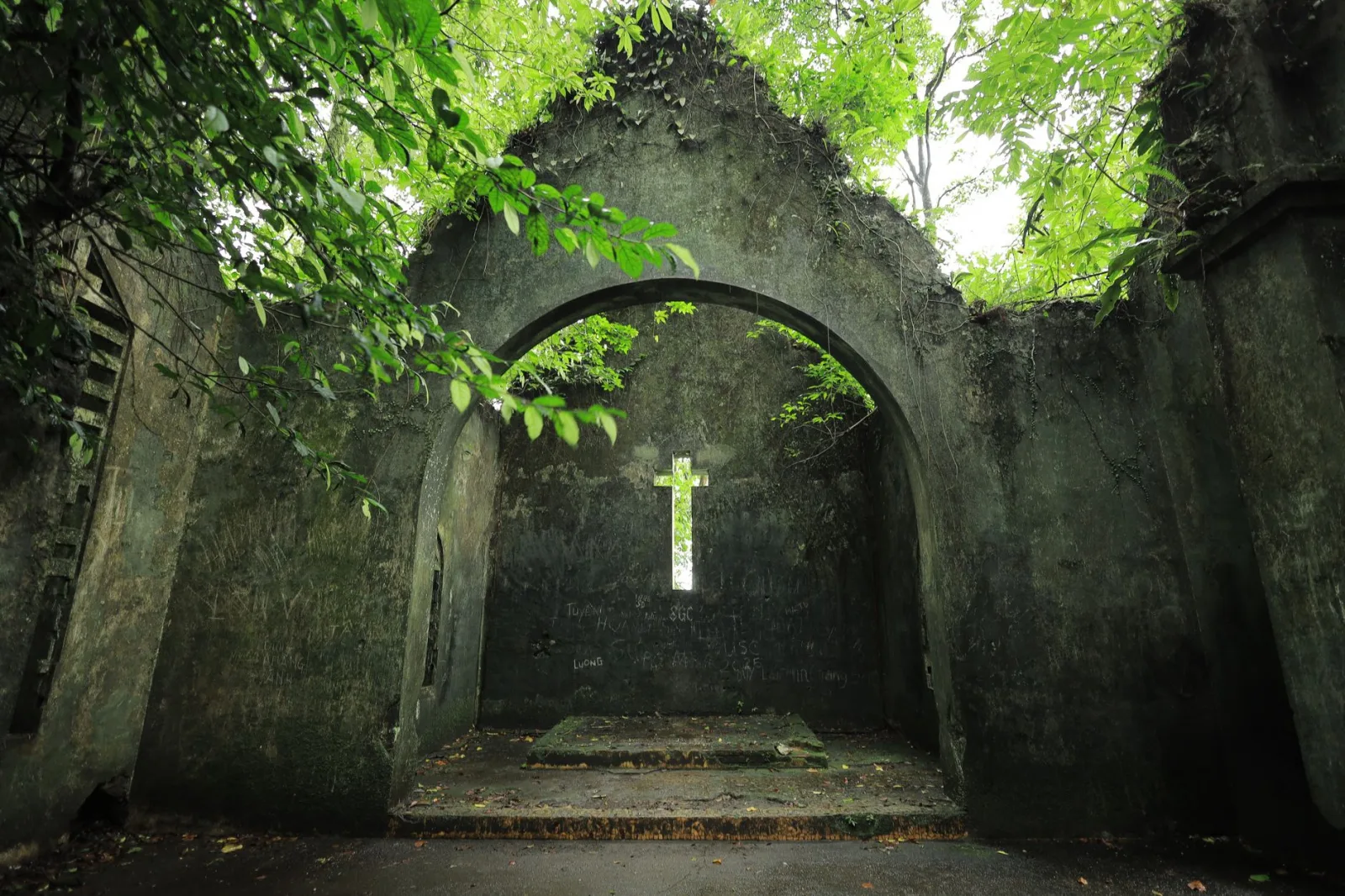 Ancient stone archway with a cross, surrounded by lush green foliage in Vietnam.