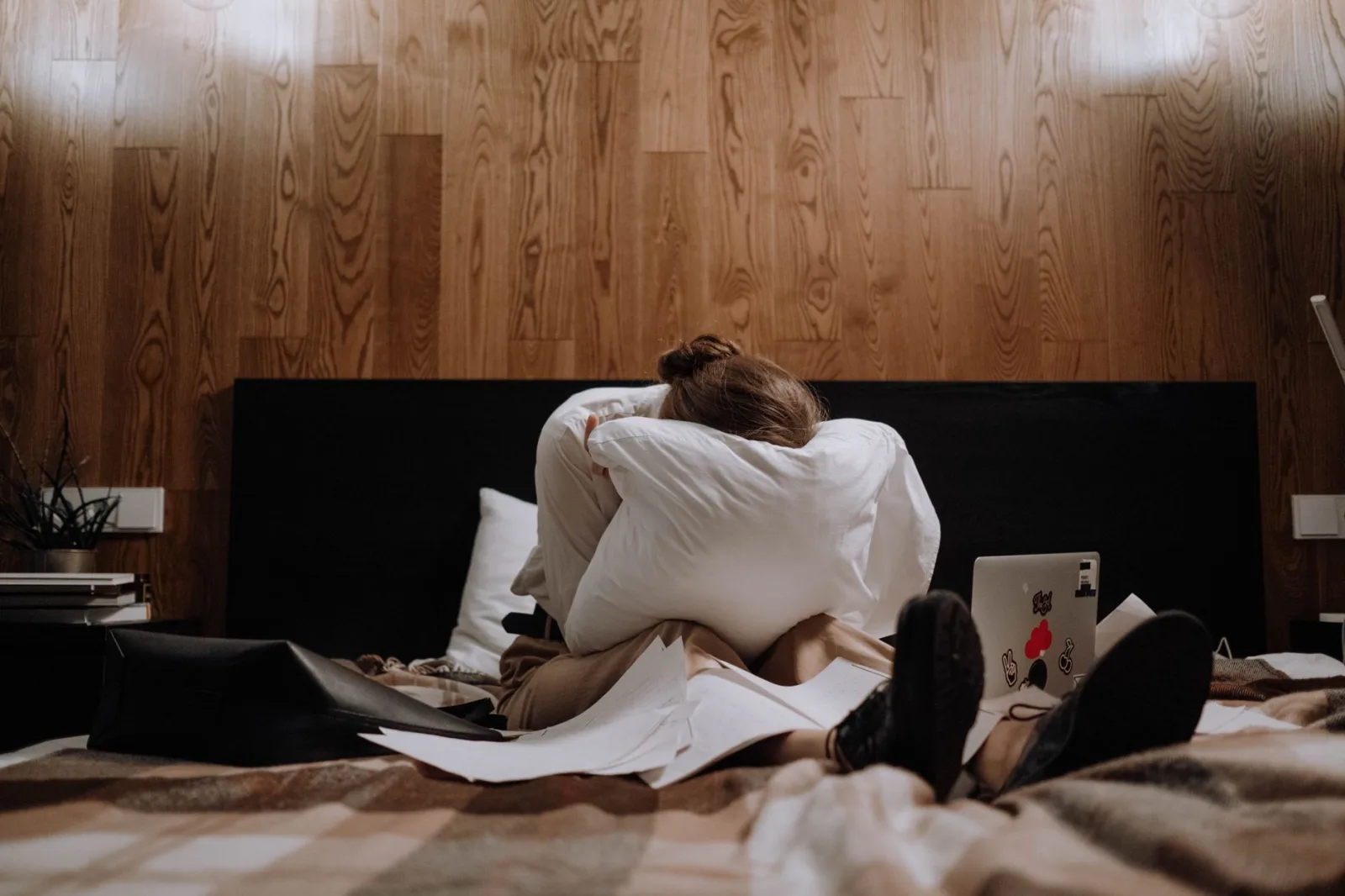 A woman sits on a bed clutching a pillow, surrounded by papers and a laptop, conveying a sense of stress.