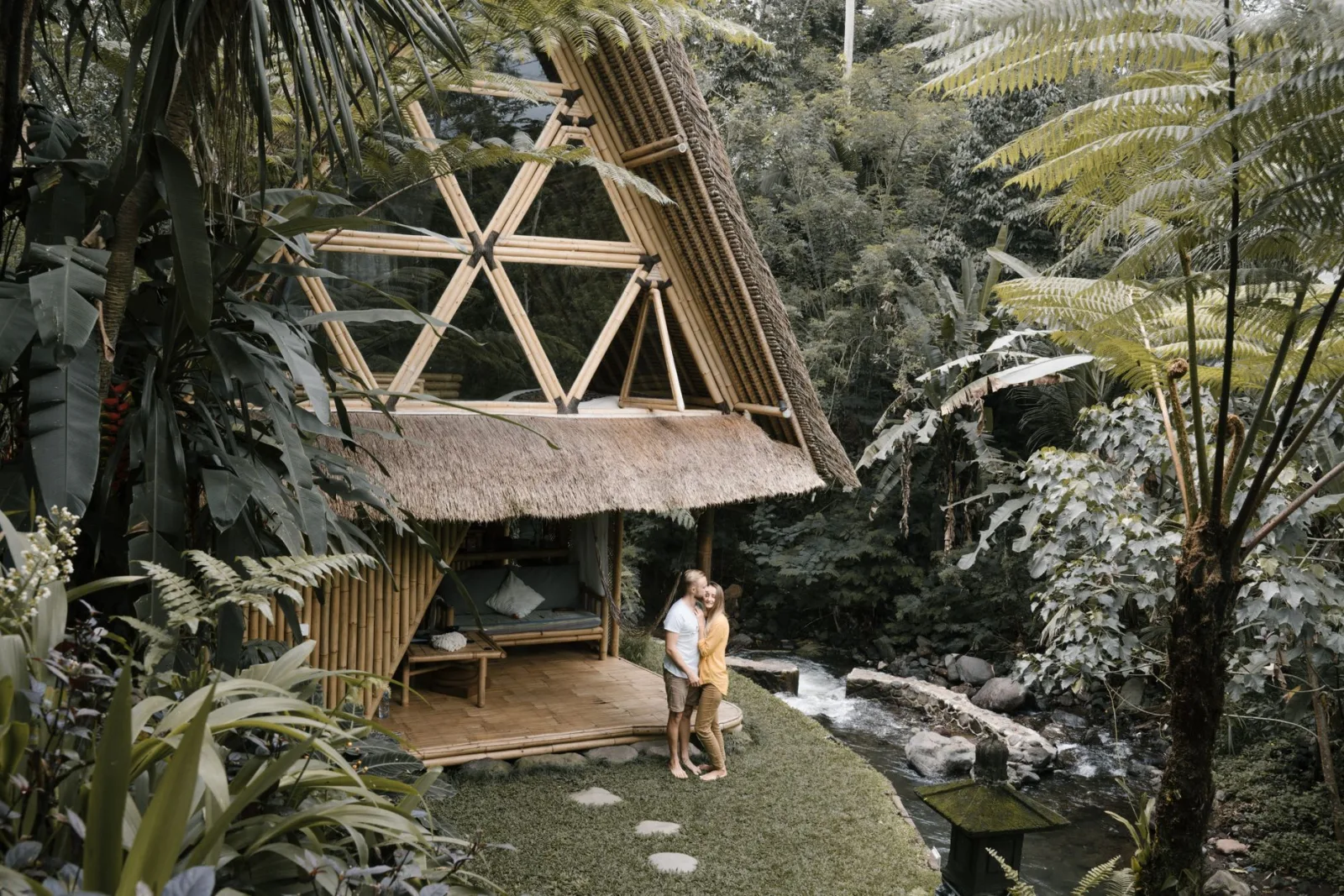 A couple embraces near a bamboo hut in a lush tropical garden by a gentle river.