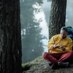 A man in a yellow jacket sits by a forest river, using a smartphone, surrounded by nature.
