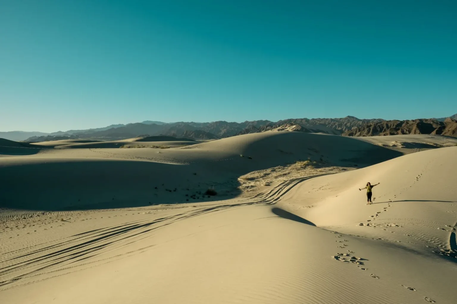 A lone traveler explores vast sunlit sand dunes under a clear blue sky.