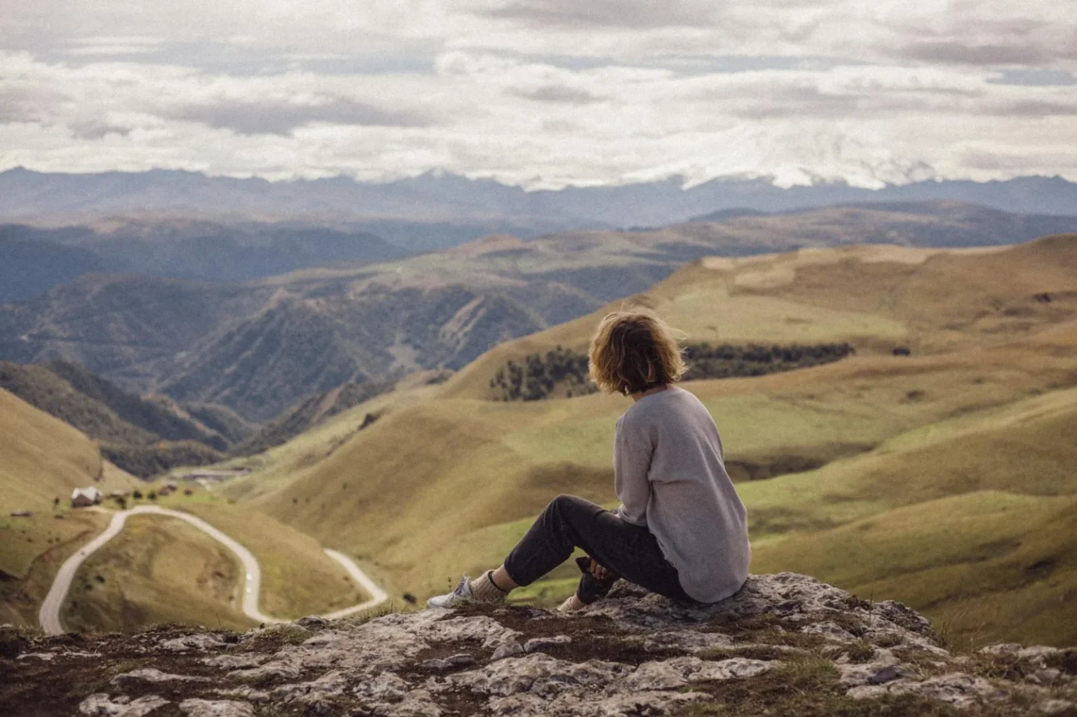 A person sitting on a hilltop, looking over a vast mountain range under a cloudy sky.