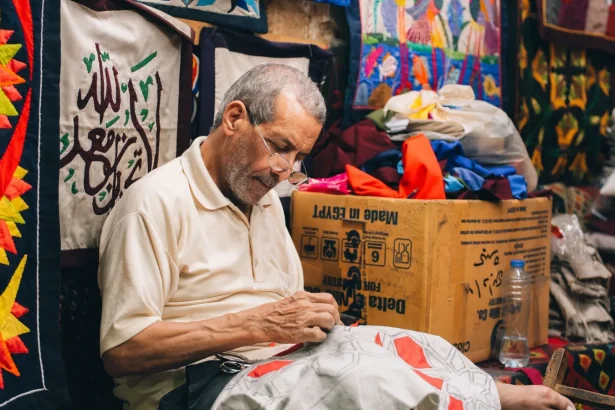 Elderly male artisan sewing traditional Egyptian textiles in a colorful workshop.