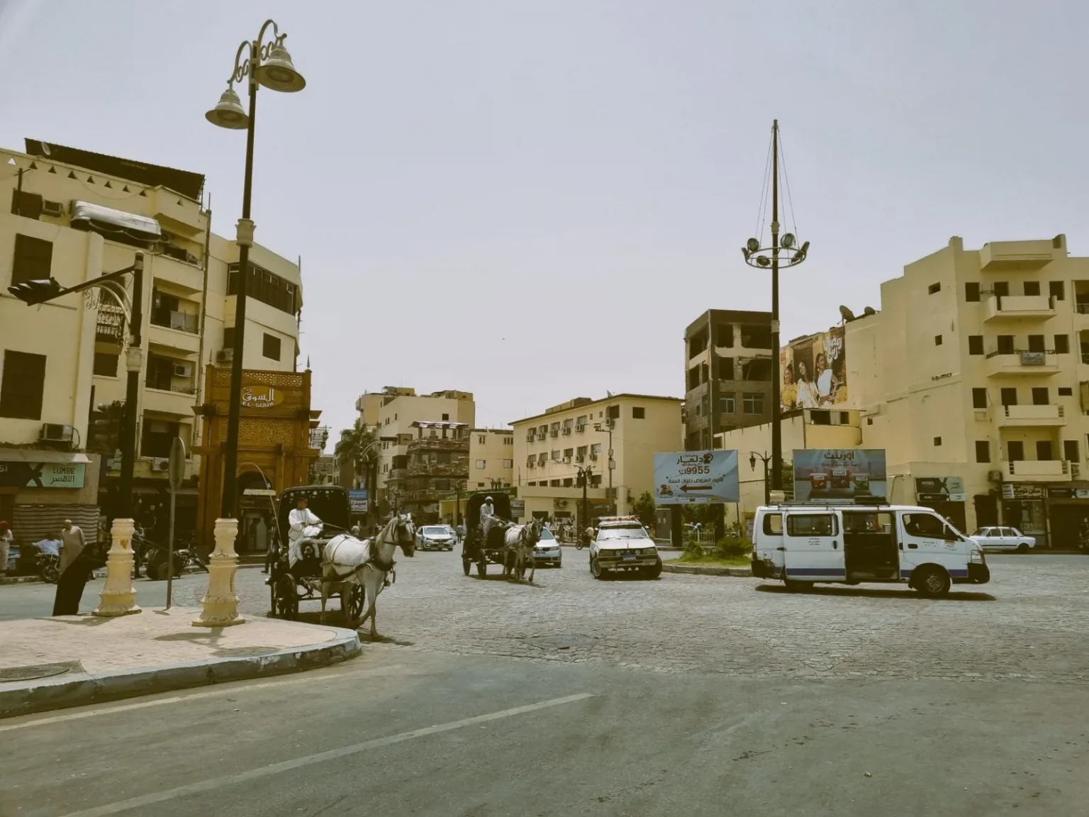 Busy intersection with horse-drawn carriages and vehicles in a Middle Eastern city.