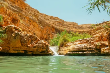 Explore a stunning oasis with a waterfall nestled in a rugged desert landscape under clear summer skies.
