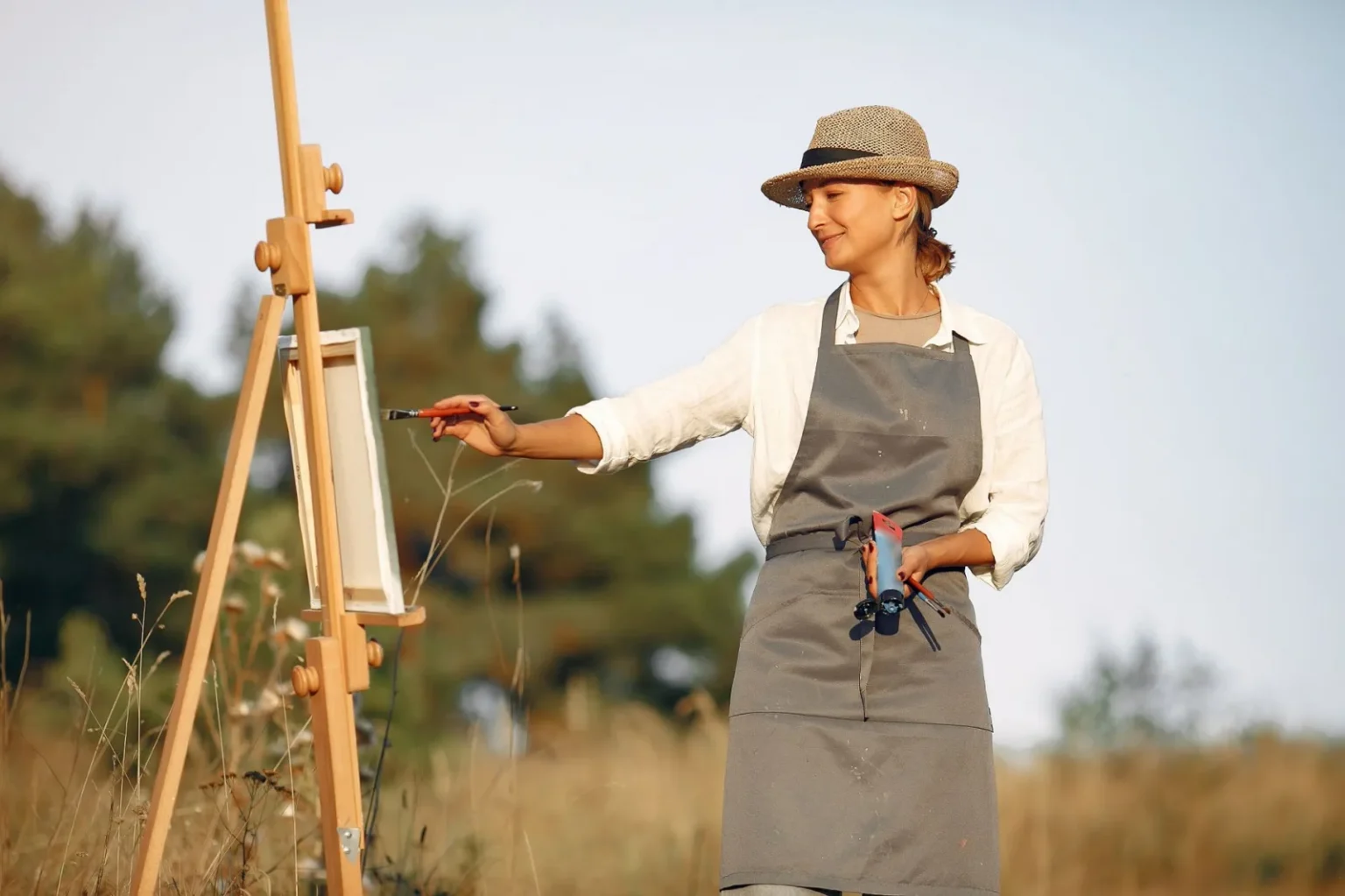 Cheerful woman in hat and apron painting while standing in summer sunny day on middle of field