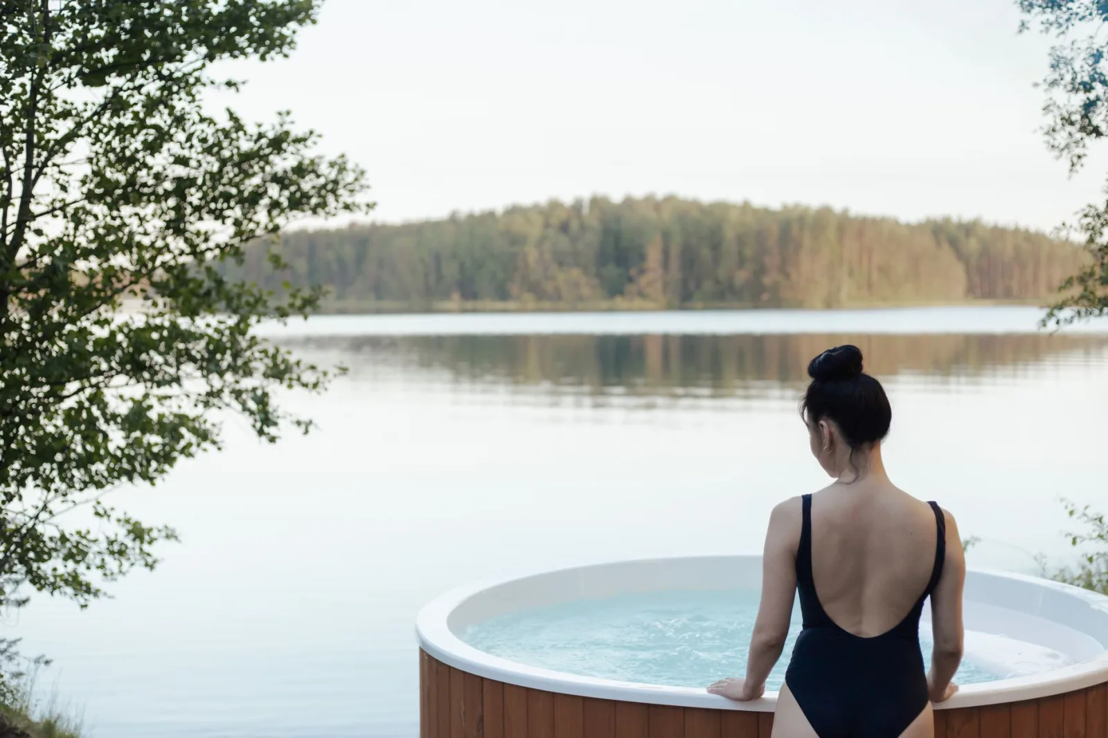 A woman in a swimsuit relaxes in an outdoor hot tub by a serene lake surrounded by trees.
