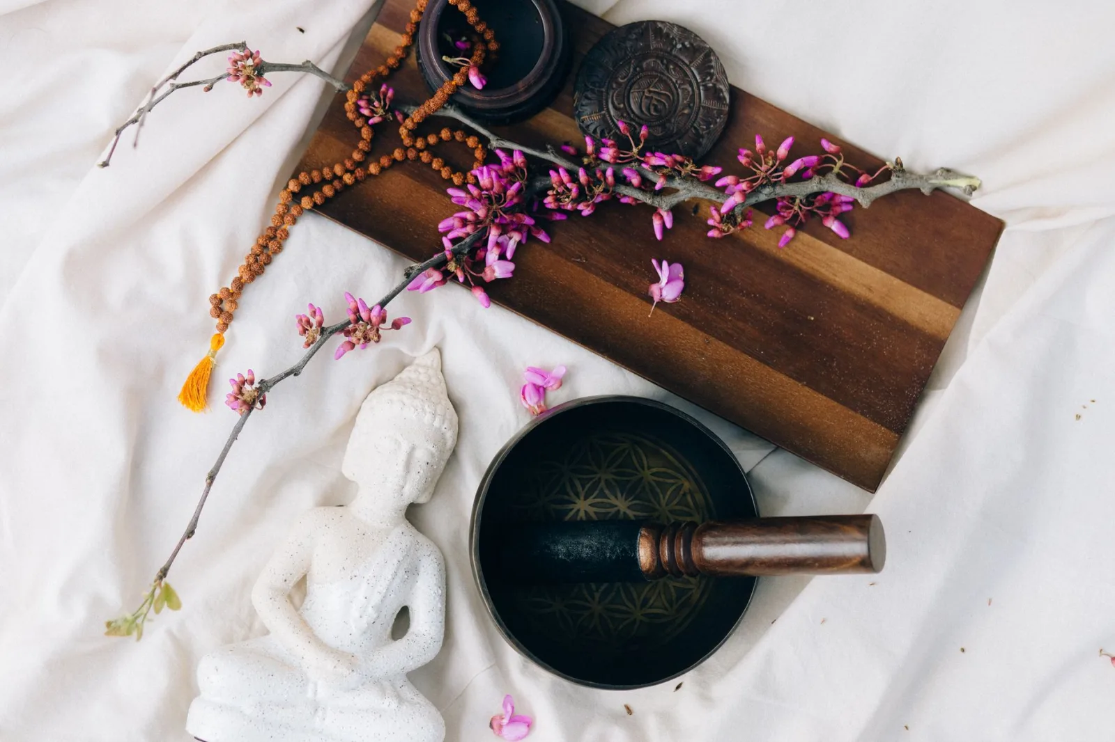 A serene Buddhist meditation setup featuring a white Buddha figurine, pink flowers, and a Tibetan singing bowl.
