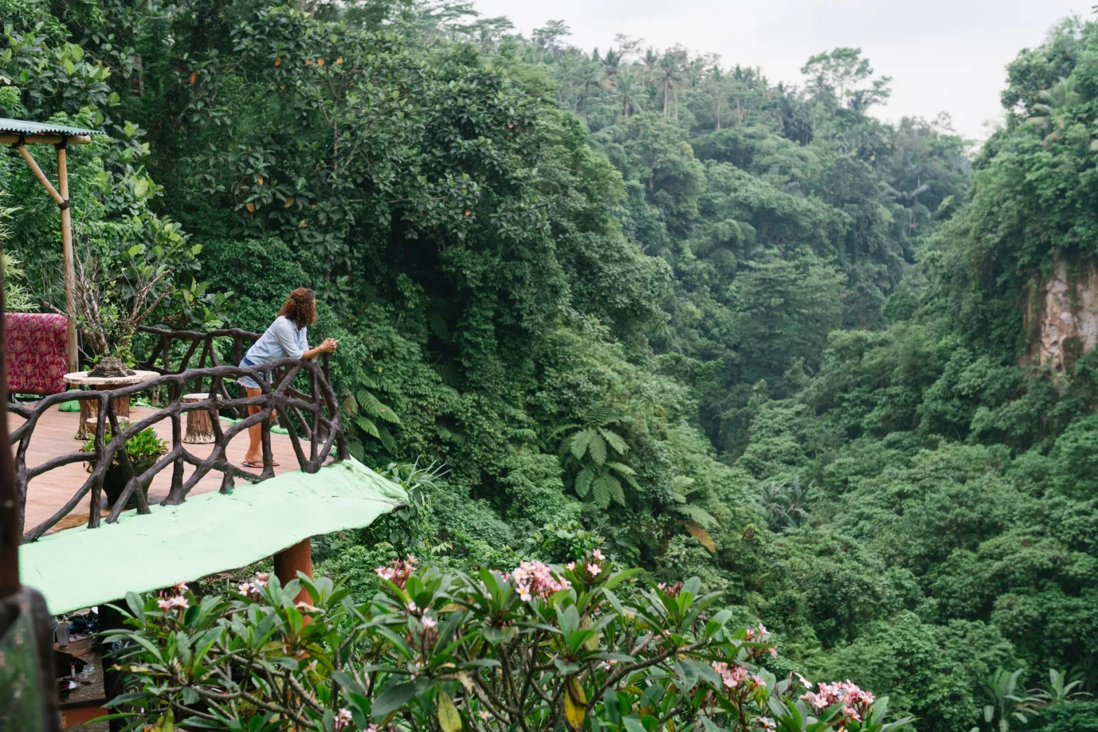 Side view full body female in denim leaning on railing on cozy rural cottage porch and admiring tropical rainforest on sunny clear day