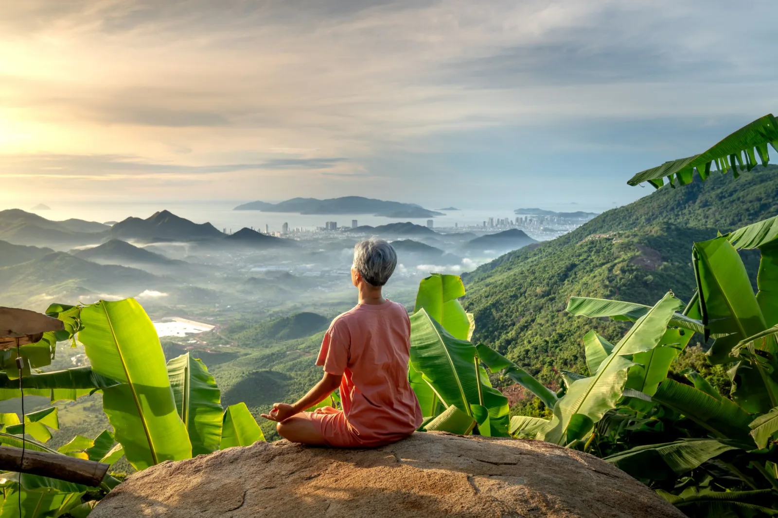 Woman meditating on rock overlooking lush green mountains and city at dawn.