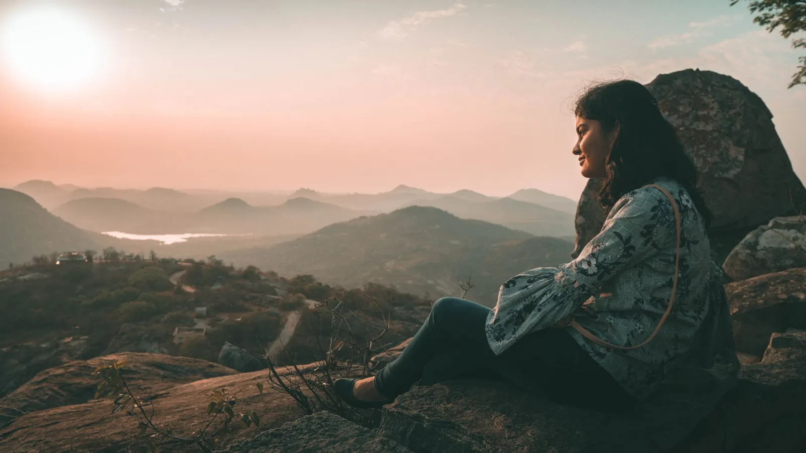 Woman sitting on a rock admires a breathtaking sunset over mountain landscape.
