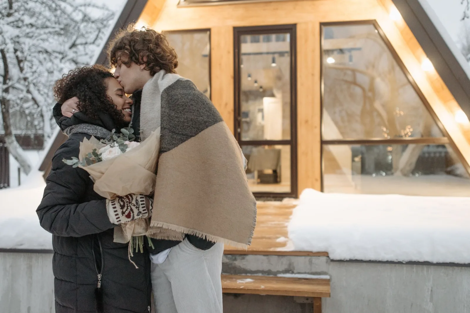 A couple sharing a heartfelt moment with a bouquet outside a snowy cabin in winter.