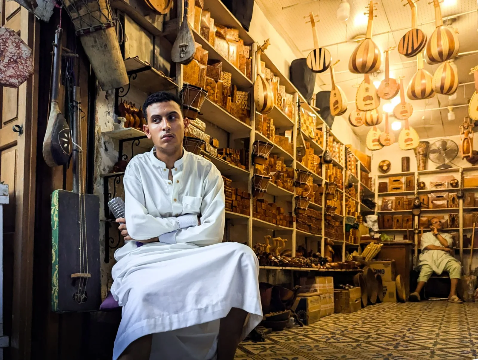 Man in traditional attire sits in a music shop filled with handcrafted lutes and instruments.