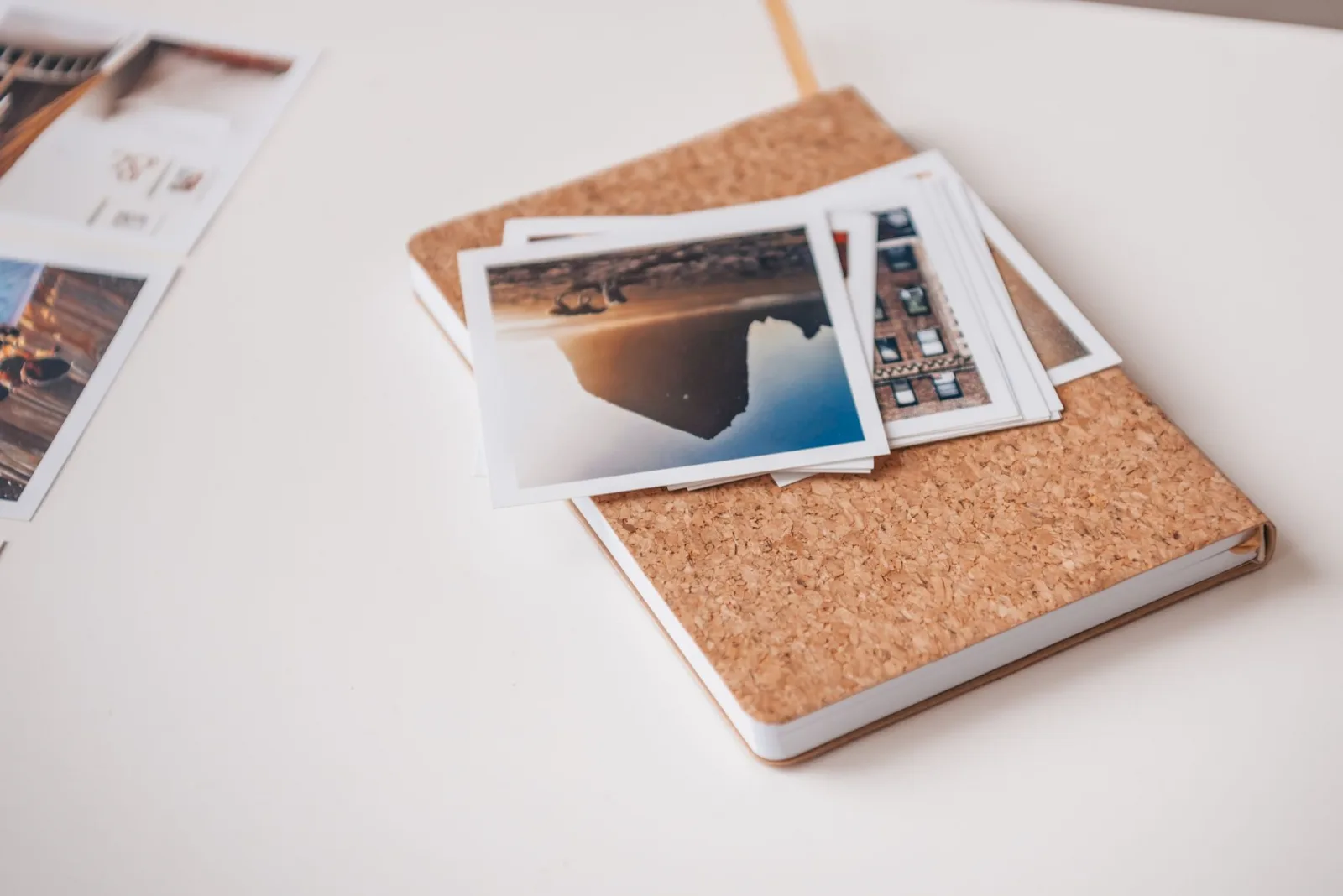 Cork-covered notebook topped with printed photographs on a white desk, conveying travel memories.