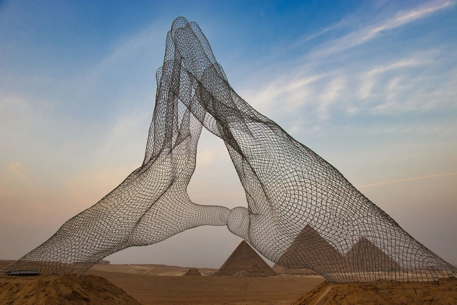 Modern wire sculpture of hands with the iconic Giza pyramids in the background at sunset.
