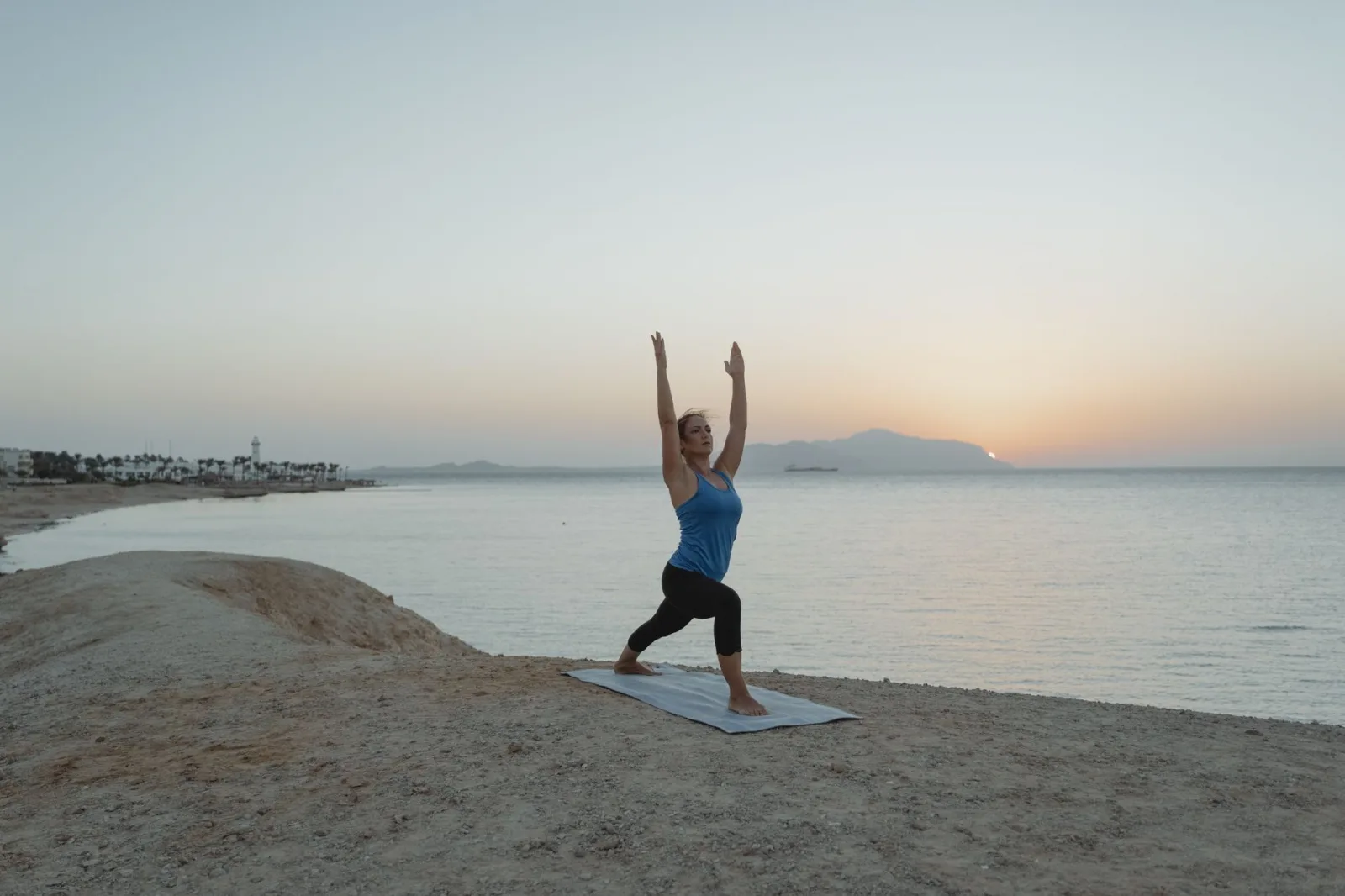 A woman practices Warrior 1 yoga pose on a beach at sunset, embracing tranquility and balance.