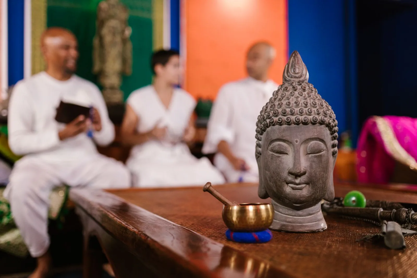 Serene Buddha head figurine on a wooden table, foreground to three meditating adults in white clothing.