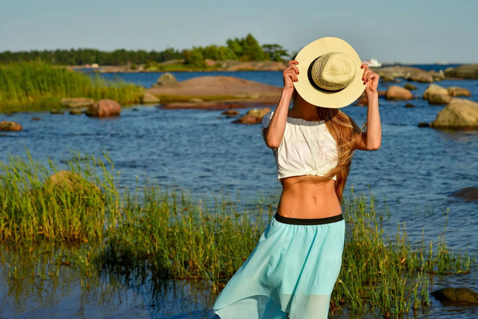Woman enjoys a summer day by the rocky beach in Helsinki, hiding face with a straw hat