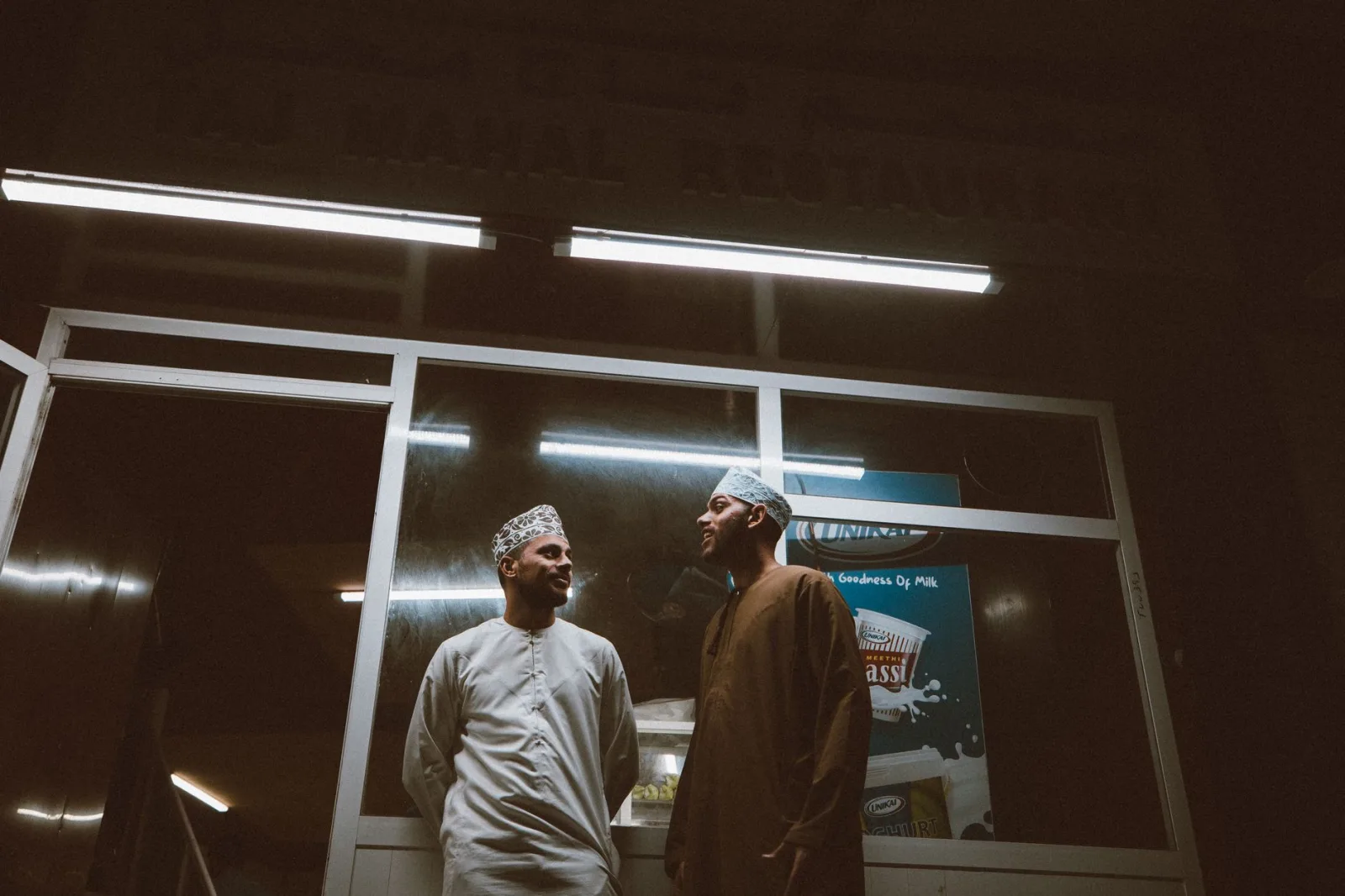 Two men in traditional Omani attire share a moment outside a Muscat restaurant at night.